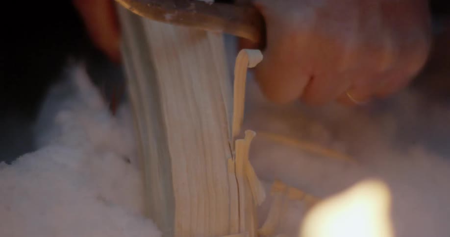Man carves a piece of wood next to a fire
