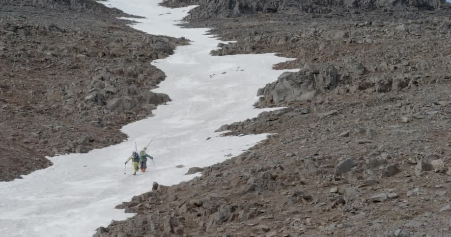 Two people carry their skis as they climb up a snow trail in the rocks