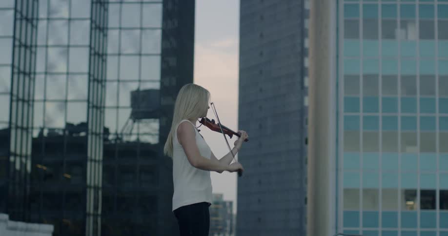 A woman plays the violin outside with skyscrapers behind her