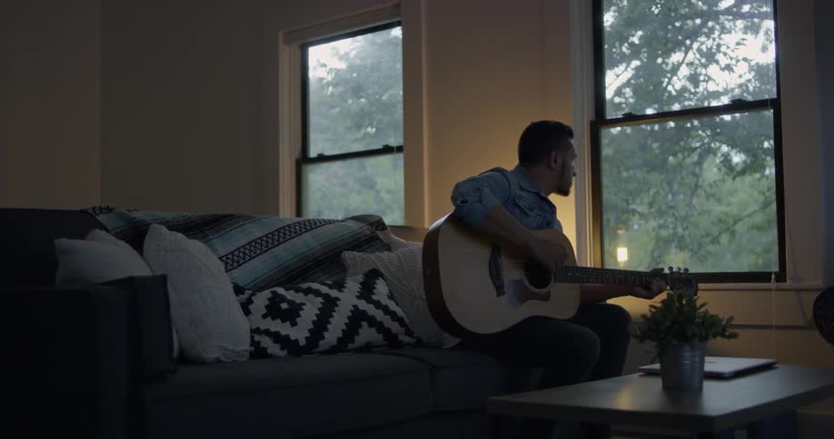 A man sits down on the couch and plays the guitar