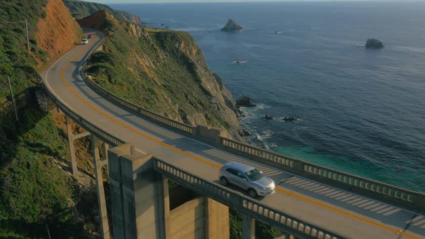 Car traveling over tall bridge on the coast overlooking the ocean as sun sets