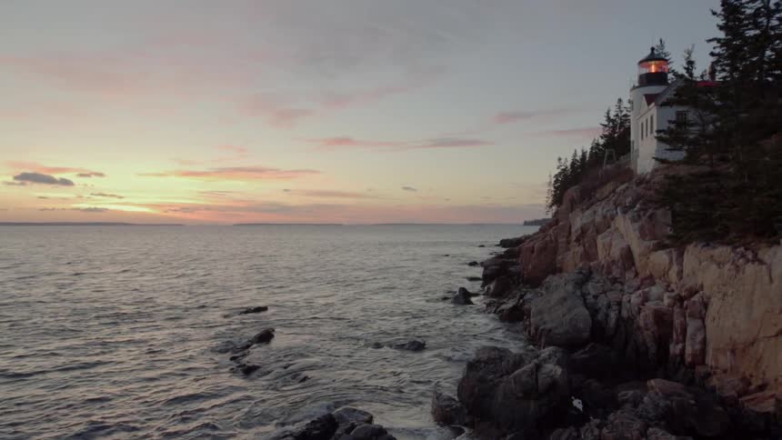 A lighthouse light glows on an ocean cliff at sunset