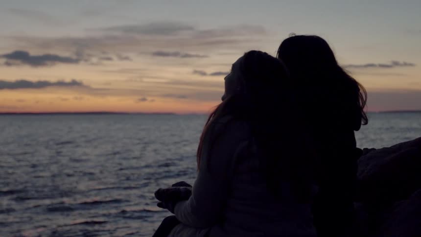 Two Women Sit By The Ocean At Sunset