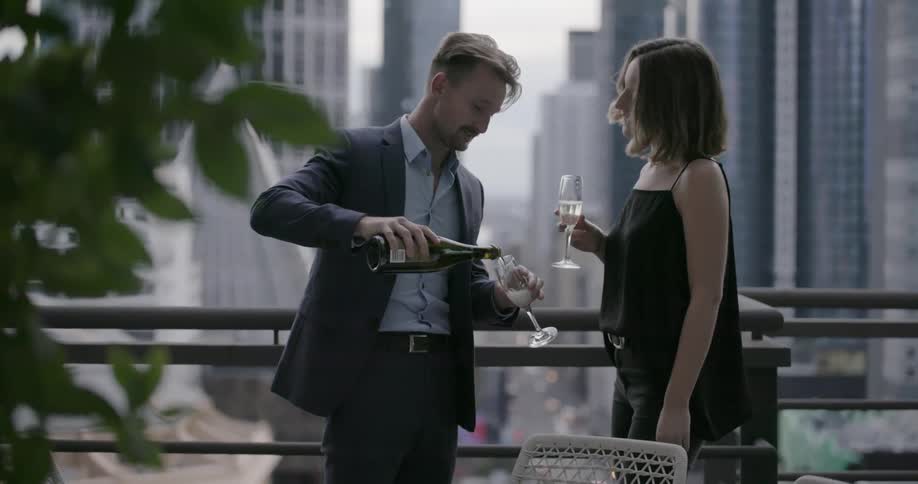 A man and woman drink champagne on a city balcony