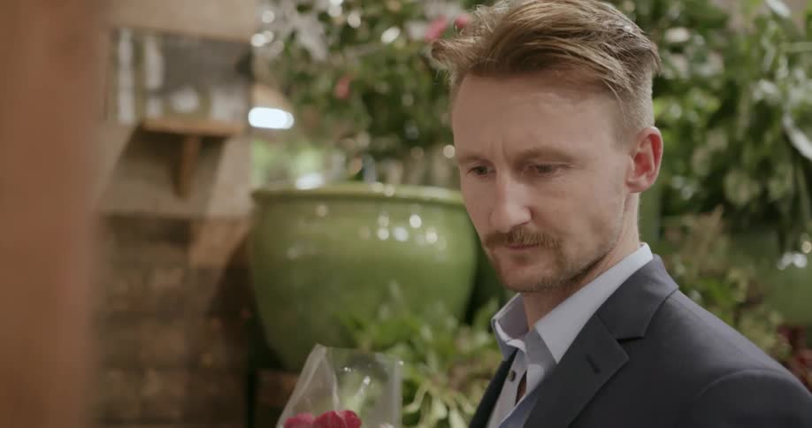 Man wearing suit and holding flower bouquet in gift shop picks up a greeting card