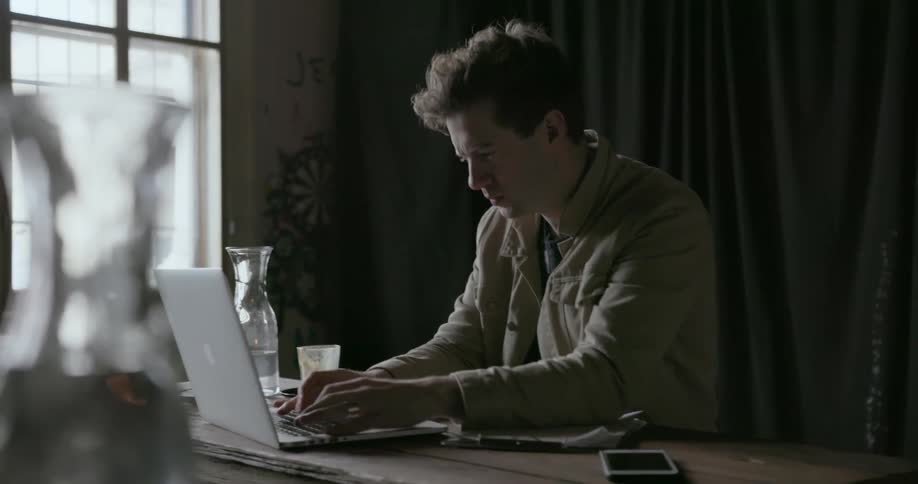 A man drinks from a cup and types on a laptop