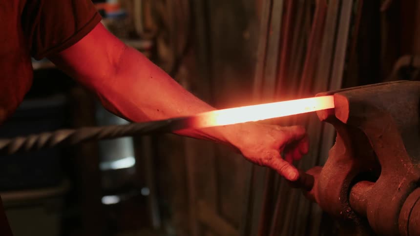 A blacksmith holds a steel rod glowing from heat with a vise