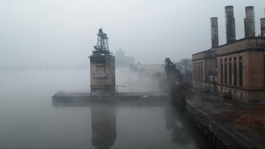 An industrial park sits alongside a body of water as fog flows onto shore