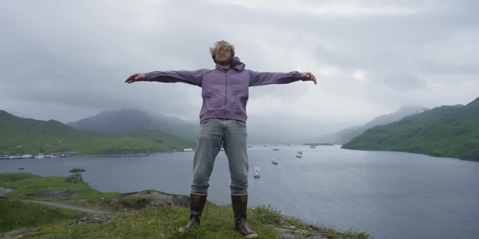 A man is standing on the edge of a cliff with his arms wide open and wind is blowing him around