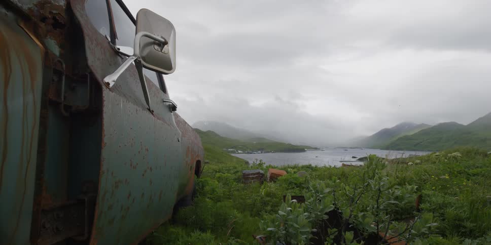 An old truck sits abandoned in a field near a lake on a cloudy day