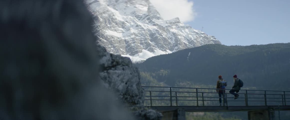 Two people look out towards mountains from a walking bridge