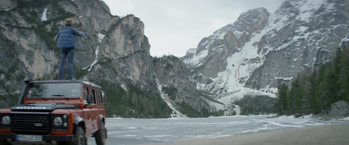 A man stands on top of a vehicle while looking at an icy mountain lake