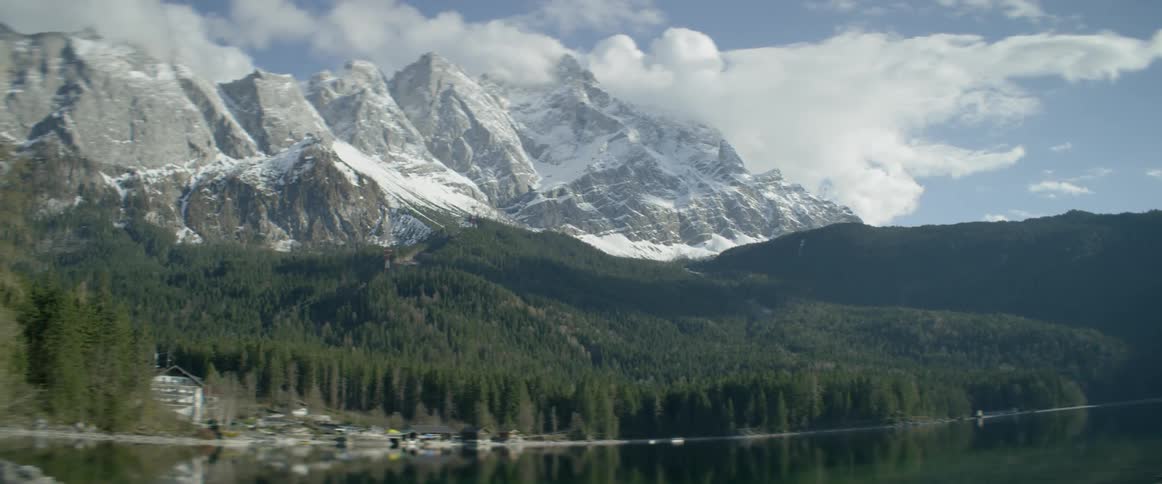 Mountains partially covered in snow in front of forest and lake on cloudy day