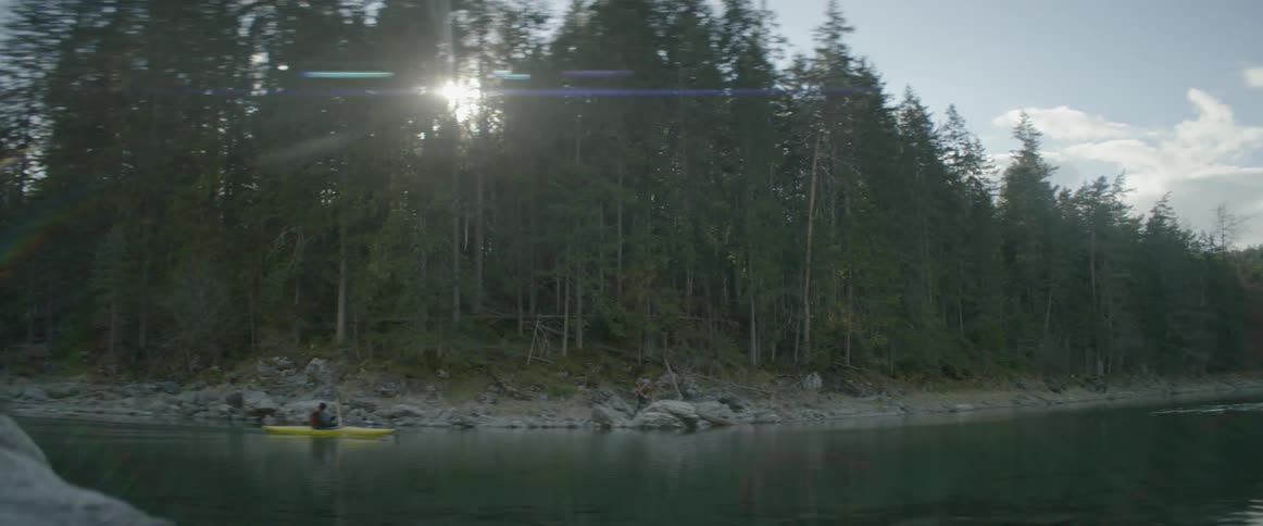 Person paddling kayak on lake as sunlight shines through forest trees
