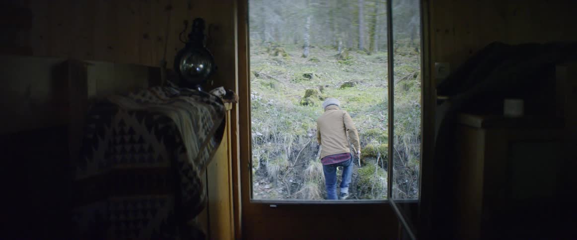 A man walks outside of a cabin in the forest while carrying an ax