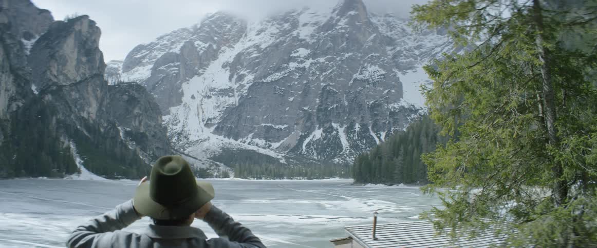 Man looking out at frozen lake and snowy mountains with binoculars