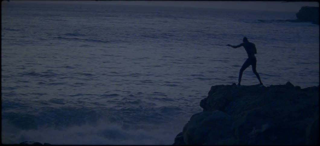A man shadow boxes on a rock near the ocean at sunset