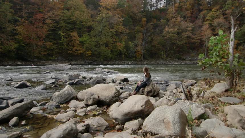 A woman sits on a rock next to a river