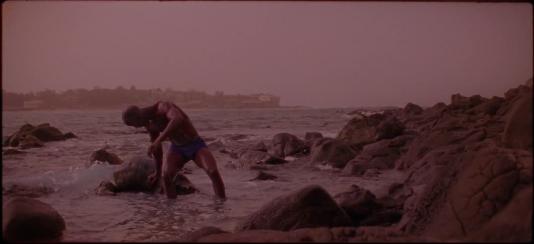 A man sits down on a rock in the ocean at sunset