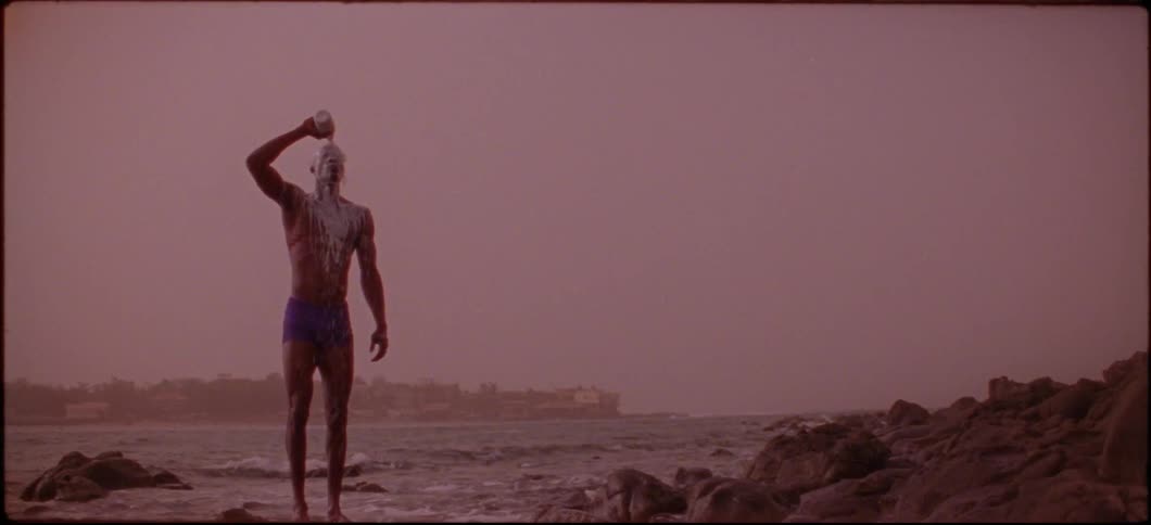 A man is pouring white liquid from a bottle and down his body with the ocean behind him