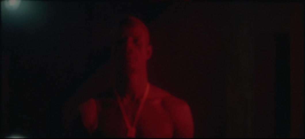 A shirtless man with a bandage on his shoulder stands outside at night