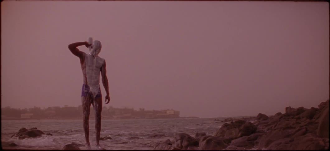 A man is pouring a white liquid on his face while standing on a beach