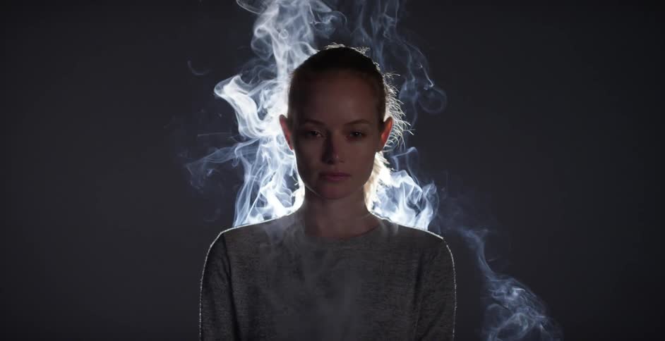 A woman stands in a dark room while being lit from behind and smoke is rising around her