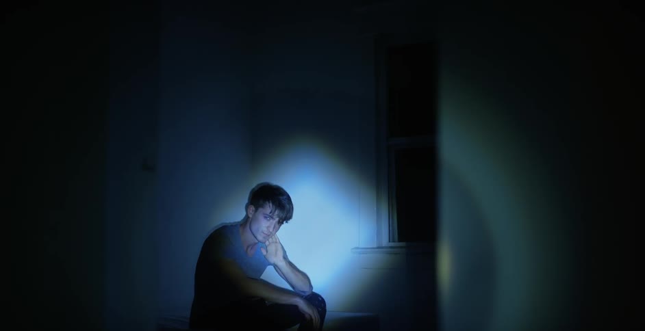 A young man sits alone in a darkened room with a flashlight pointing at him
