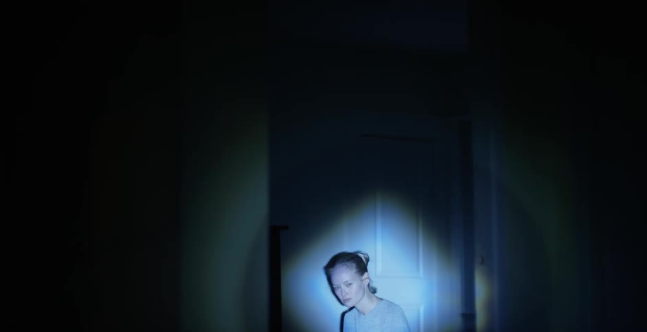A woman sits alone in a dark room with a flashlight pointing at her, and then looks toward the light