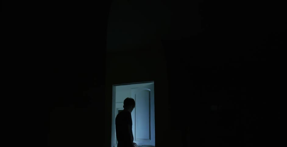 A man standing in total darkness opens a door into a lighted room