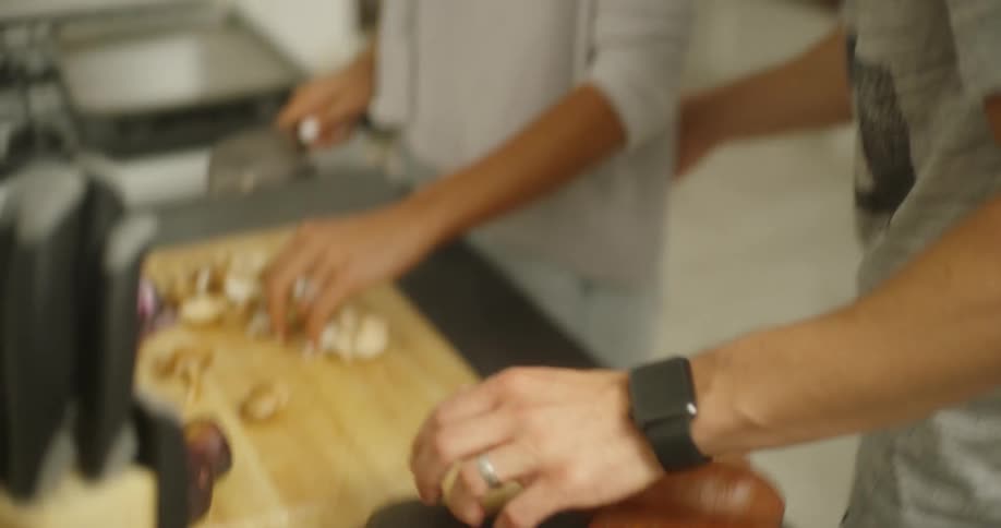A man brings a handful of yams to a woman as she slices mushrooms in their kitchen