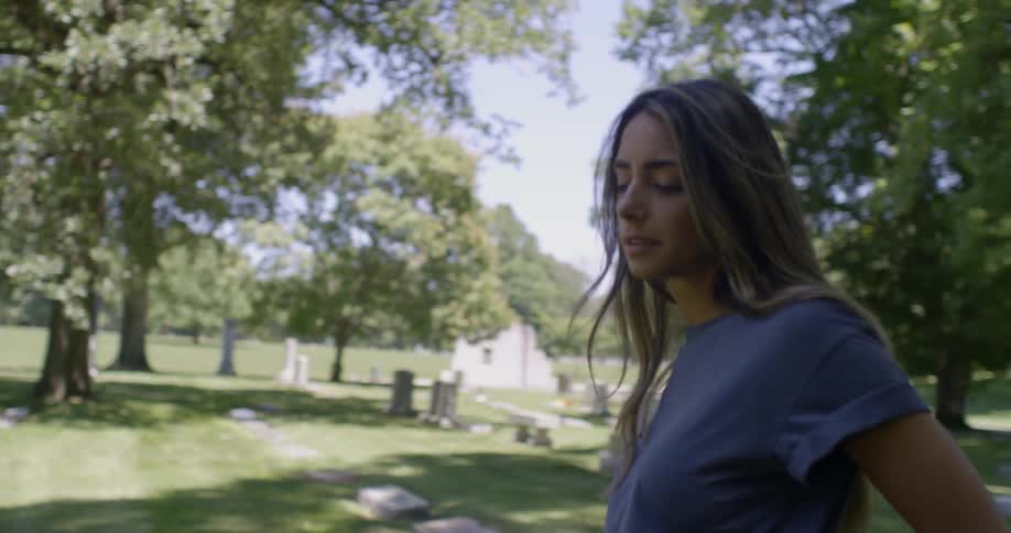 A woman is standing in a graveyard looking down at a tombstone