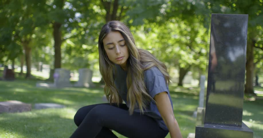 A woman is sitting in front of a tombstone in a graveyard