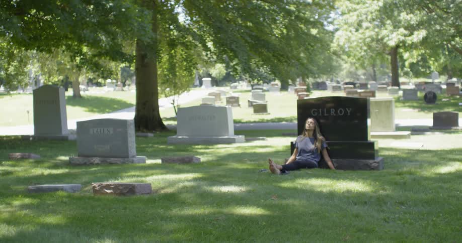 A woman is sitting with her back against a headstone in a cemetery during the day