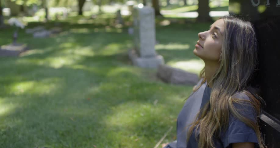 A woman leans against a headstone in a cemetery
