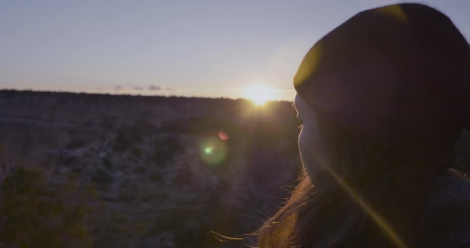 A view of a young woman looking into the distance as the sun sets behind cliffs