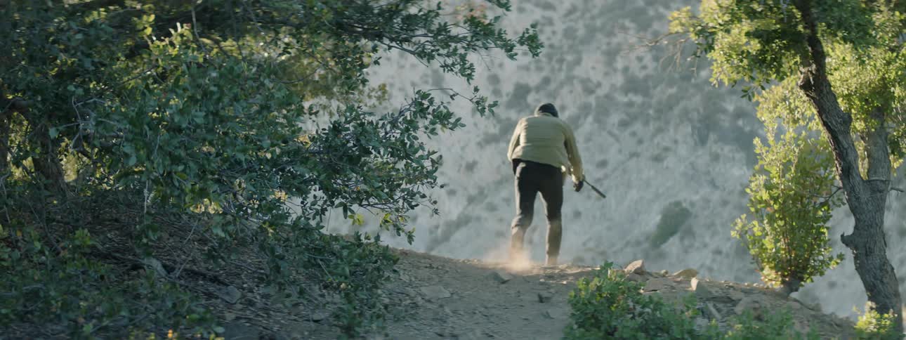 A man holding a rifle and kneeling on a dirt path gets up and moves away