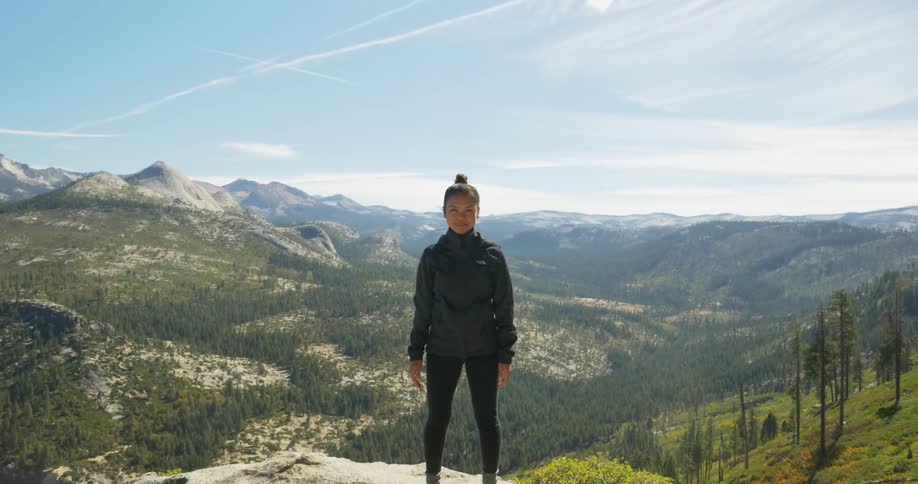 A woman stands a top a tall mountain with a small grin