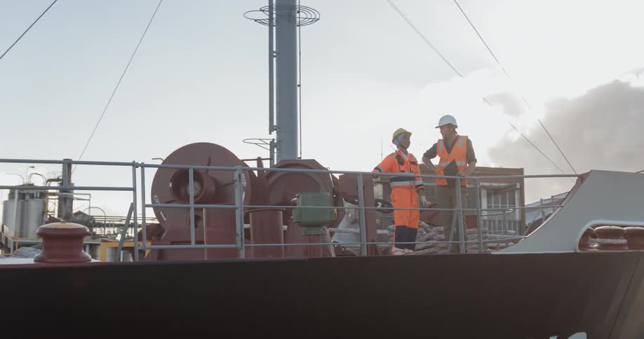 Two men wearing vests and hard hats lean on the railing of a ship