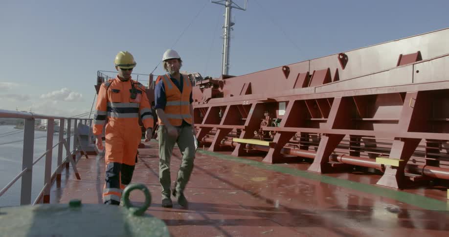 Two people in safety gear are walking along the deck of a ship