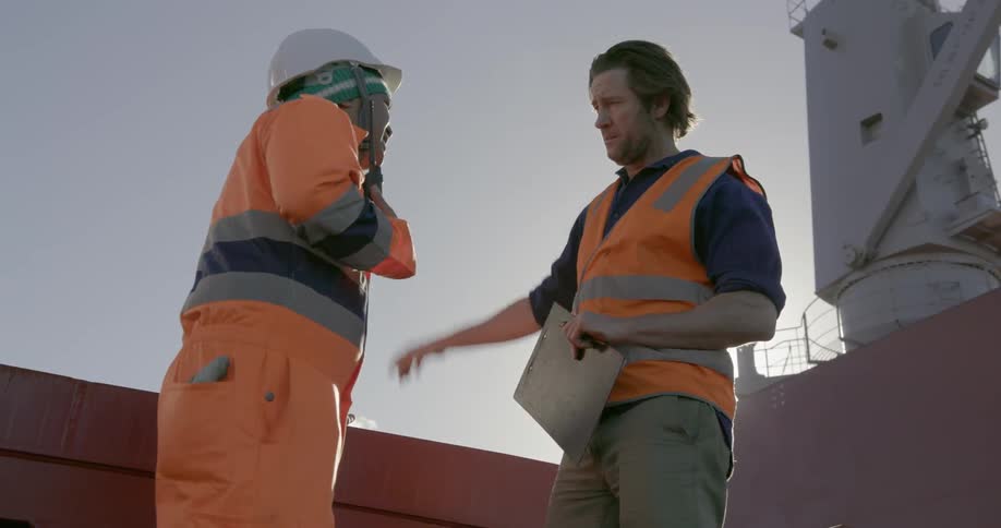 Two people wearing safety gear are talking on the deck of a ship