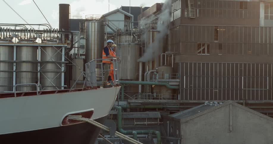 Two men wearing vests and hard hats lean on the railing of a ship