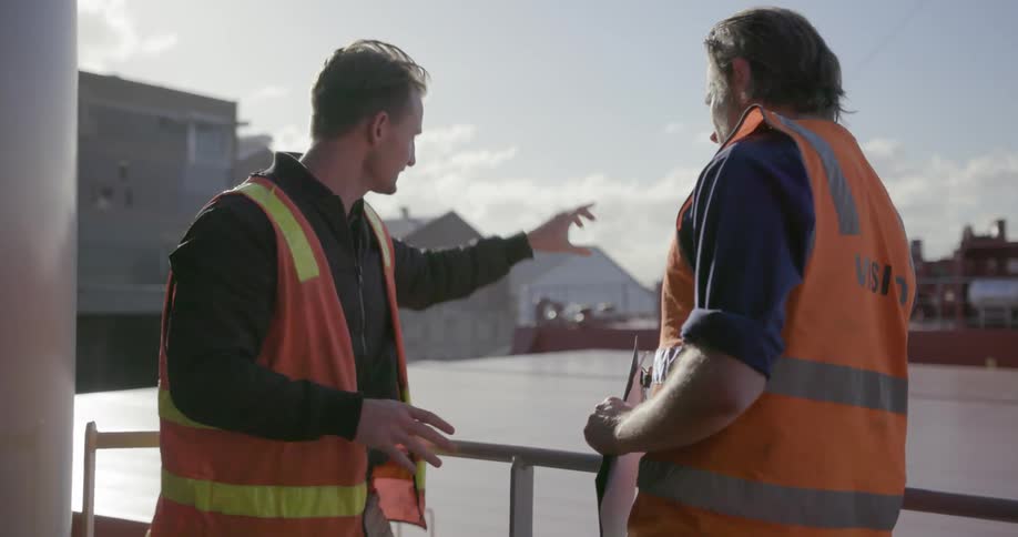 Two people in safety vests are leaning against a railing discussing something