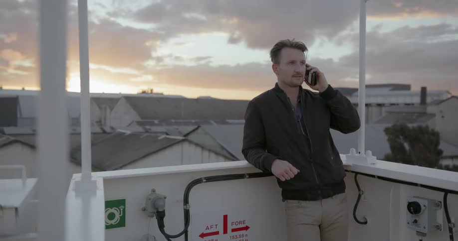 A man leans on a rail while talking on the phone at sunset