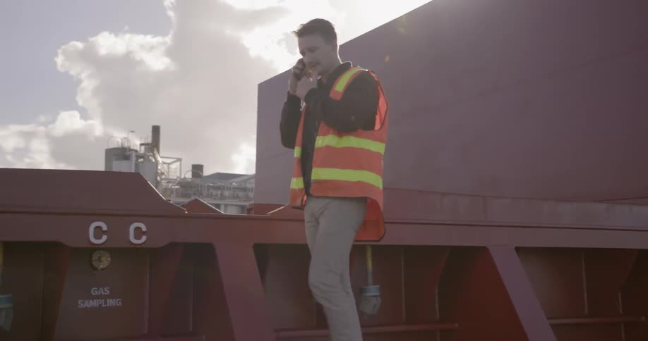 Man wearing an orange vest walks in an industrial area talking on the phone