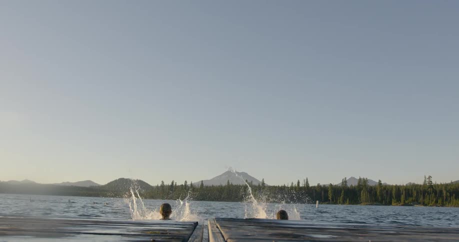Two girls leap off a pier into a lake on a sunny day