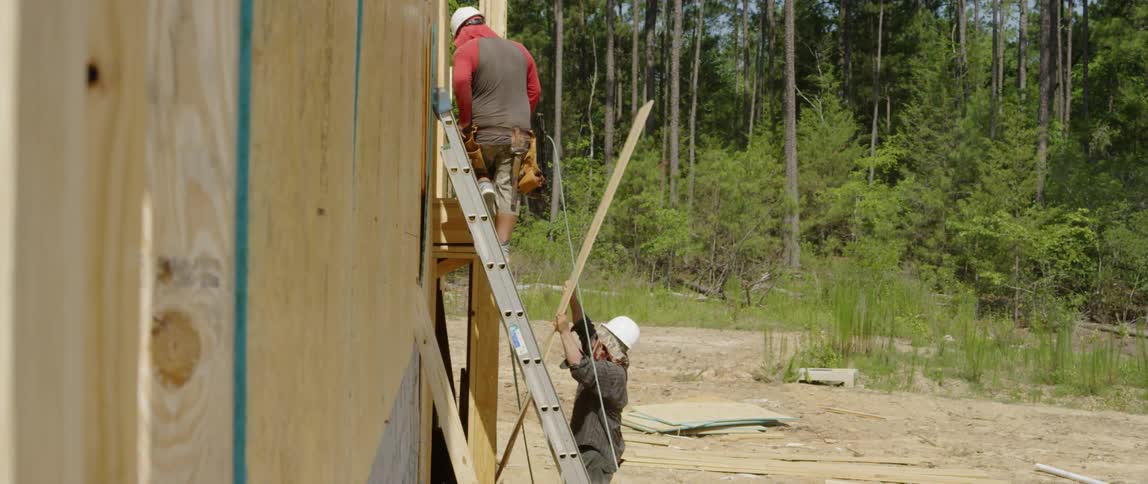 A construction worker carries a wood board to another worker on a ladder