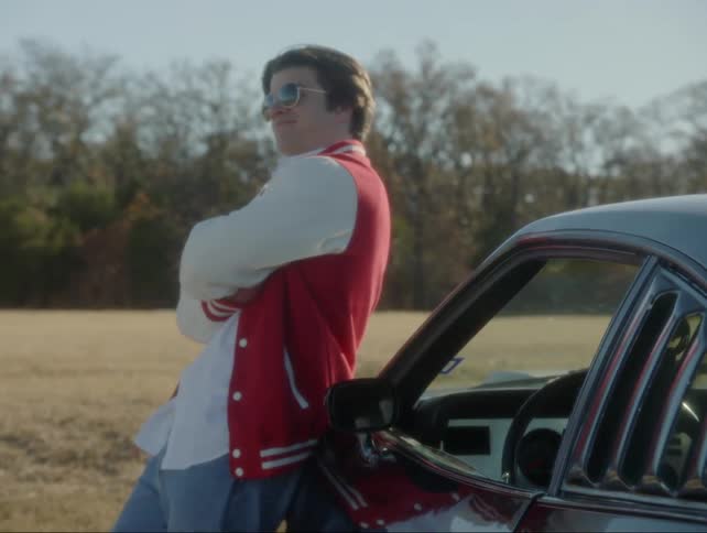 A boy in a varsity jacket leans against his car in the sunshine