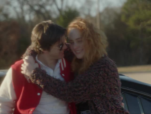A teenage couple are hugging and smiling as they stand outside during the day