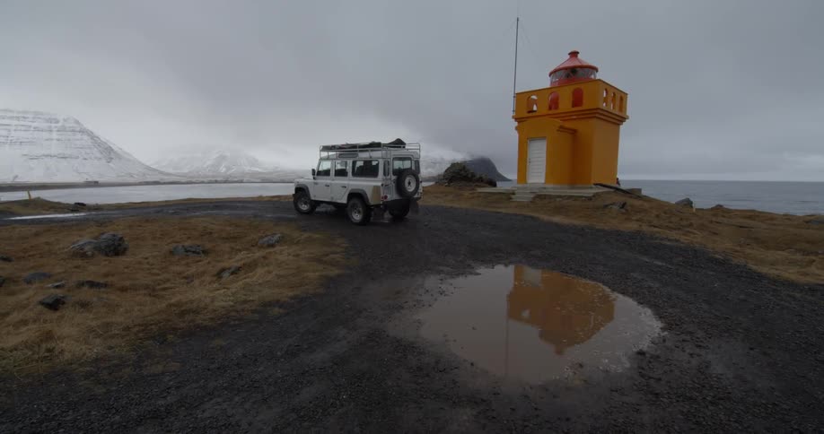 A white truck sits parked by a lighthouse near the water and mountains